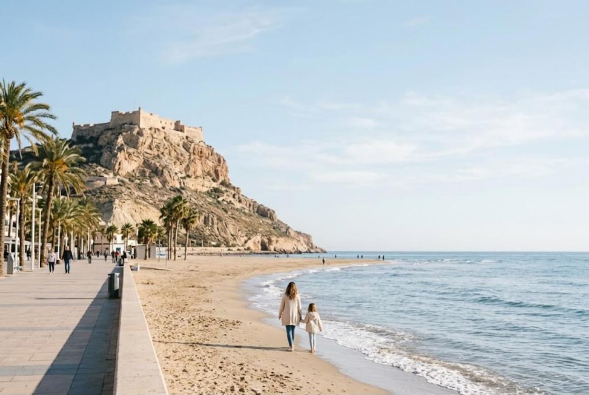 Madre e hija de la mano por la orilla de una playa de arena junto a un paseo marítimo con palmeras. Al fondo un castillo corona un monte rocoso junto al mar
