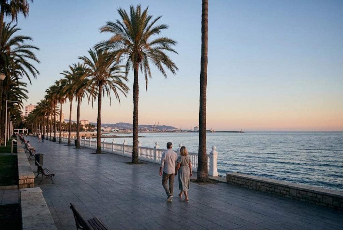 Pareja paseando al atardecer por un paseo marítimo con palmeras y barandilla blanca junto al mar. Al fondo se ven el puerto y los montes