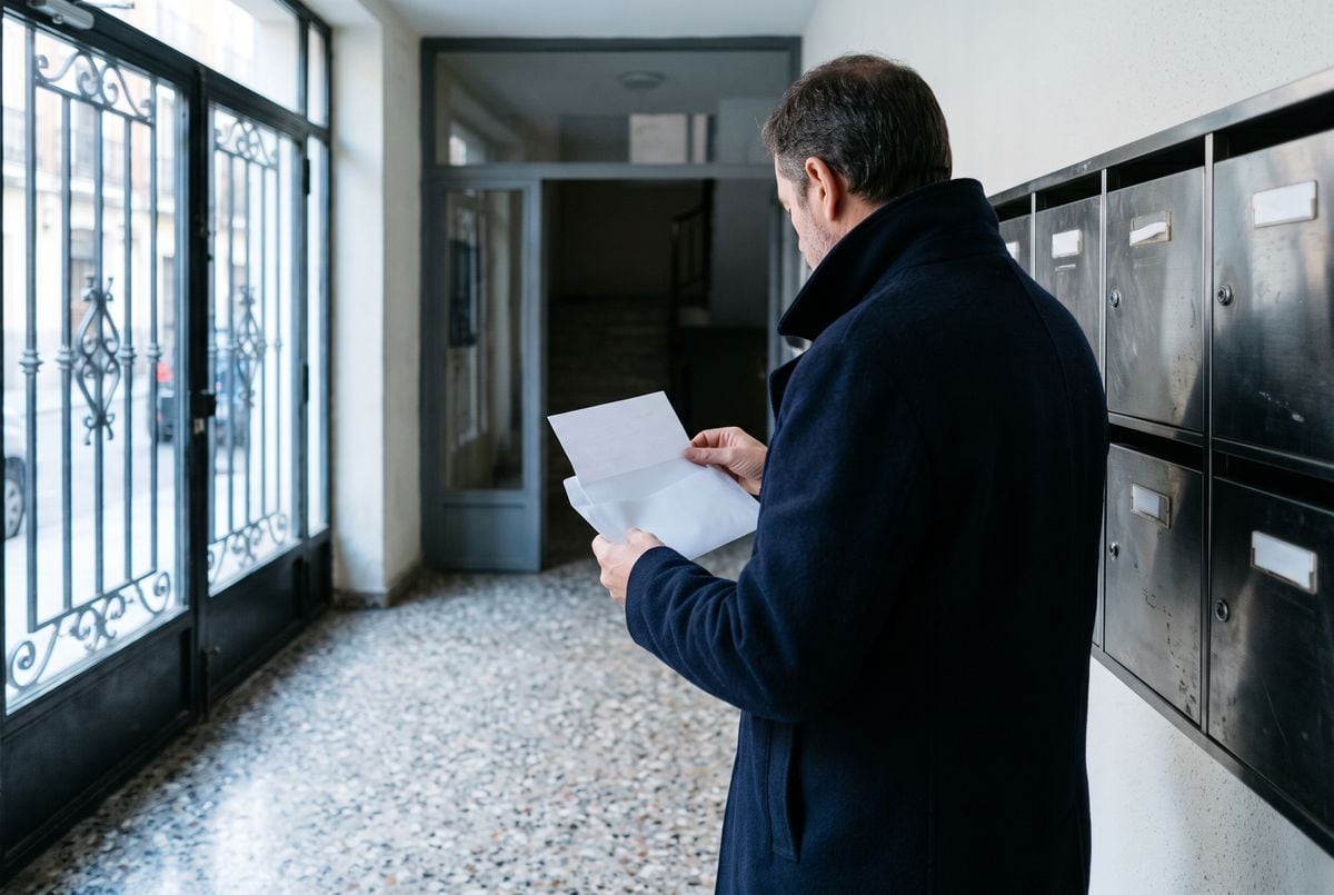 Hombre de espaldas abriendo un sobre junto a los buzones del portal de su edificio en una mañana de otoño
